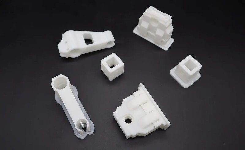 A collection of white 3D-printed parts created by the Raise3D Pro3 Plus HS. The parts, displayed on a dark surface, include complex geometric shapes and mechanical components, demonstrating the printer’s precision and ability to produce intricate designs. The samples showcase varying heights, widths, and detailed features, emphasizing the printer's versatility and high-resolution printing capabilities.