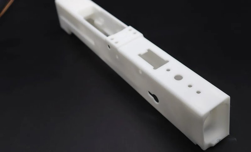 A long, rectangular, white plastic part created by the Raise3D Pro3 Plus HS 3D printer. The object features multiple cutouts and holes of varying shapes and sizes, likely designed for specific industrial applications or assembly purposes. It rests on a dark surface, highlighting its smooth texture and precision engineering.