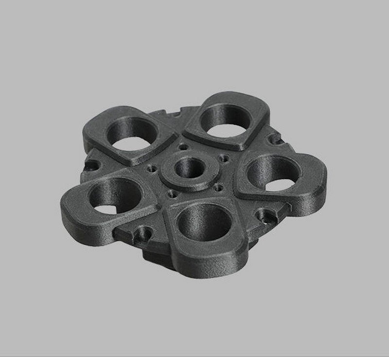 Black component part created by the Raise3D Pro3 Plus HS, featuring a complex structure with multiple circular openings and a central hub. The piece demonstrates the printer’s ability to produce detailed, functional prototypes or end-use parts. The object rests on a neutral gray background, highlighting its high-quality finish and precise details.