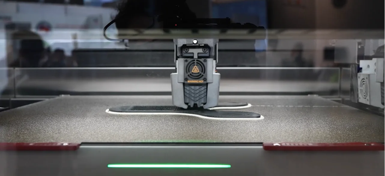 Raise3D RMF500 3D printer creating a part with black and white filament on a textured surface in a workshop setting.