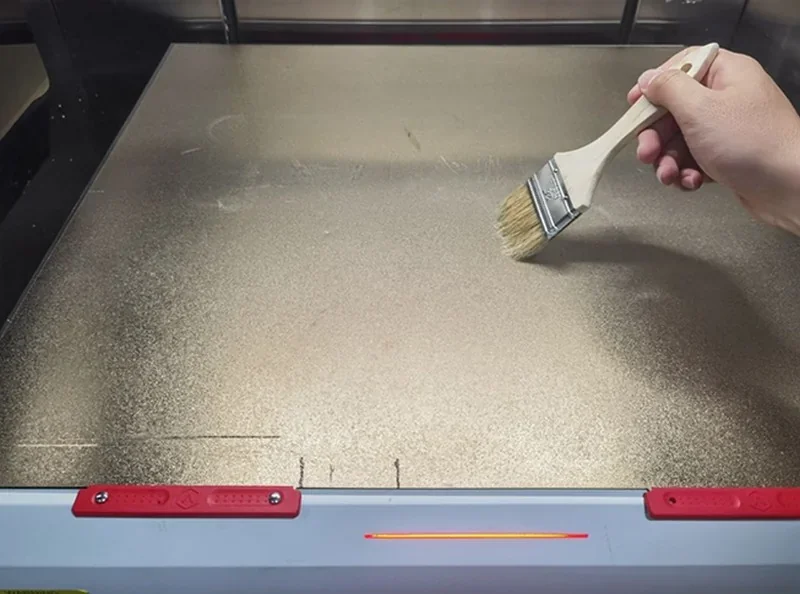 Cleaning the Raise3D RMF500 print bed with a brush, highlighting maintenance process for high-quality 3D printing.