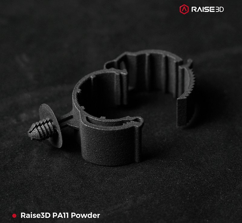 A close-up view of a black, ruggedly-textured part created by the raise3d rms220 using PA11 powder, showcasing its complex structure with multiple cylindrical openings and a screw-like attachment. The component rests on a dark surface, highlighting the contrast and precision of the printing. The Raise3D logo is visible in the corner, emphasizing the brand.