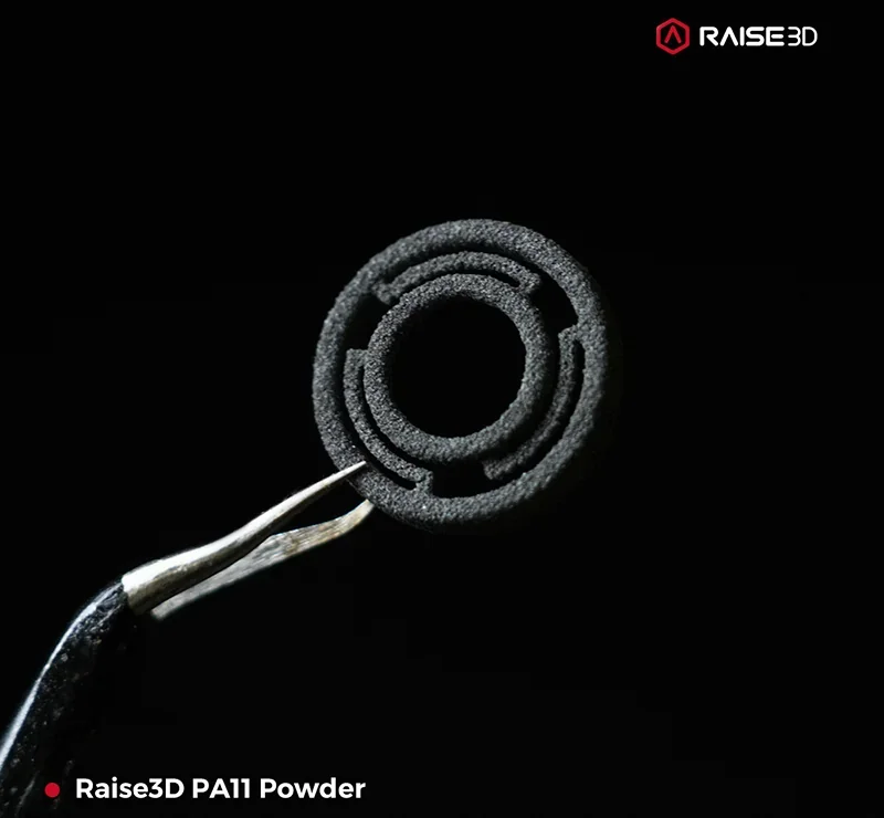 Raise3D RMS220-produced PA11 powder part held by tweezers, featuring intricate circular design against black background, with Raise3D branding.