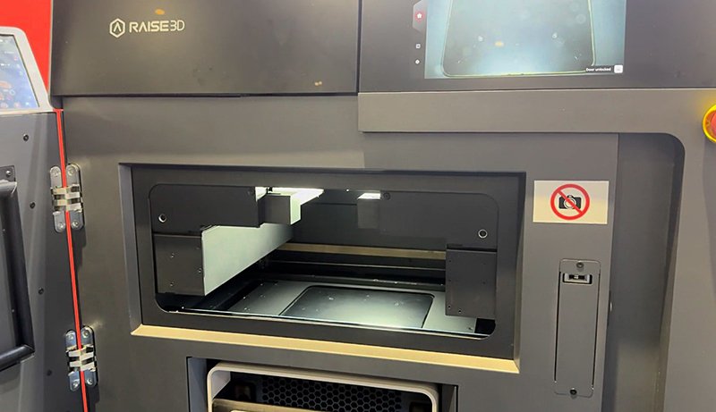 Interior view of the Raise3D RMS220 3D printer, showcasing the printing chamber. The open front door reveals the intricate components inside, designed for high precision printing tasks. The interior is illuminated, showing the platform area clearly, with a textured surface for optimal material adhesion. Visible are the dual side hinges and an 