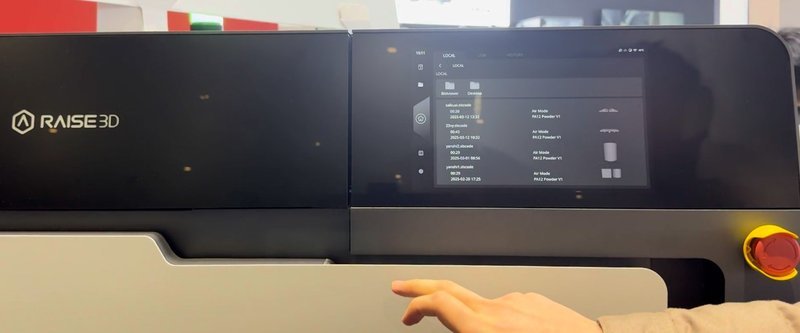 The control interface of the Raise3D RMS220 3D printer shows a full-color touchscreen displaying the printer's operational settings and job queue. The screen is positioned on the upper section of the printer, adjacent to the Raise3D branding. Below the screen, a prominent emergency stop button is visible, indicating safety features. The interface is designed for user-friendly interaction, showing current operation status and previous print jobs.
