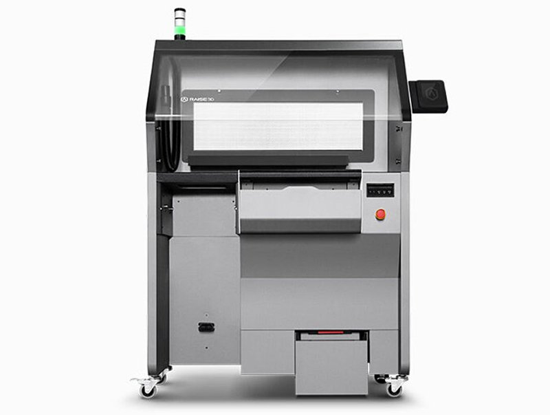 The Raise3D RMS220 3D printer in a full, front-facing view, displaying detailed features and structural components. The sleek design includes a large, transparent window that allows users to monitor the printing process. Below the window, various compartments and access panels are organized effectively. The printer is mounted on wheels for easy mobility, and branding is visible on both the top section and a display screen to the right. The industrial design features a gray and black color palette, emphasizing its professional application setting.