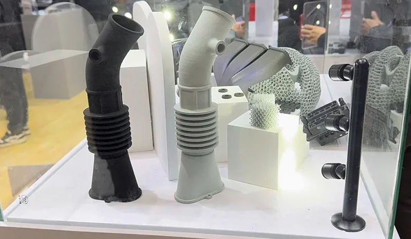 Raise3D RMS220 printed parts displayed in a case, featuring black and gray pipe models, lattice structure, and geometrical samples on white pedestals.