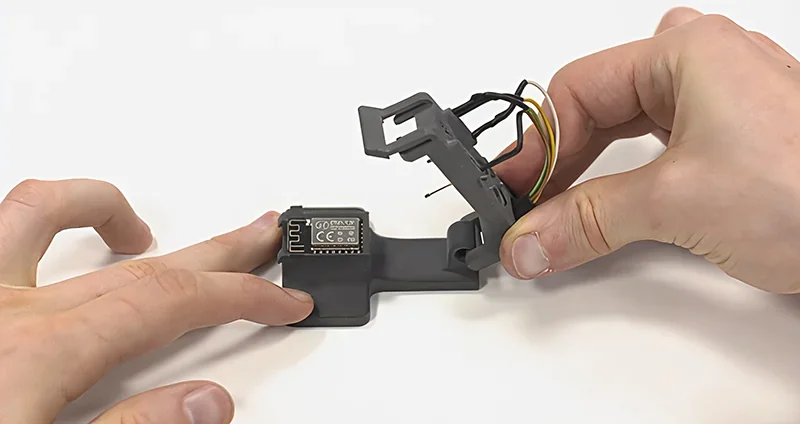 Hands hold a 3D-printed electronic mount with wires, created by Sinterit Lisa X, on a white background.