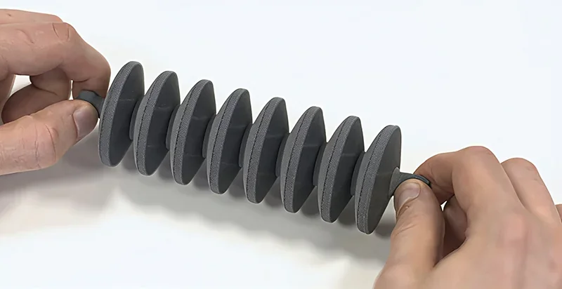 Hands holding a flexible, gray 3D-printed object made by Sinterit Lisa X on a white background.