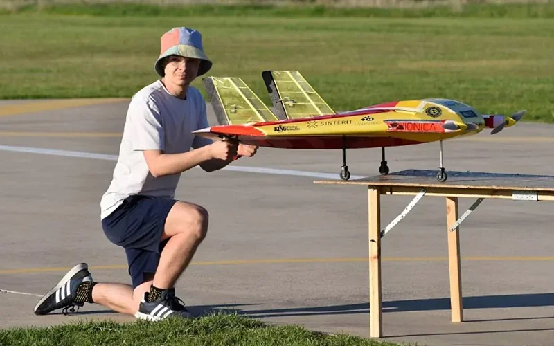 Person kneeling on pavement with yellow-red model plane, labeled "Sinterit," likely 3D printed by Sinterit Lisa X, on wooden table, grassy background.