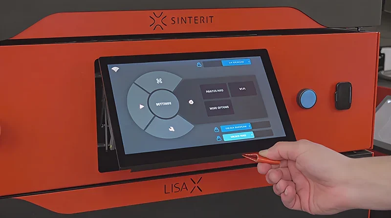 Sinterit Lisa X 3D printer with touchscreen interface, showing settings menu. Hand holding a sample part, likely created by the printer.