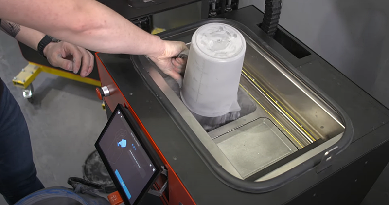 Sinterit Lisa X 3D printer with a hand removing a printed part from the chamber; touchscreen interface visible; industrial setting.