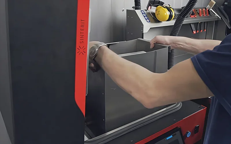 Sinterit Lisa X 3D printer with operator loading material, branded red panel, control screen visible, tools and earmuffs on adjacent shelf, industrial setting.