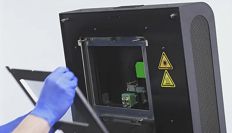 Sinterit Lisa X open chamber, with a person in blue gloves, symbols indicating laser and heat, designed for creating precise 3D printed parts.
