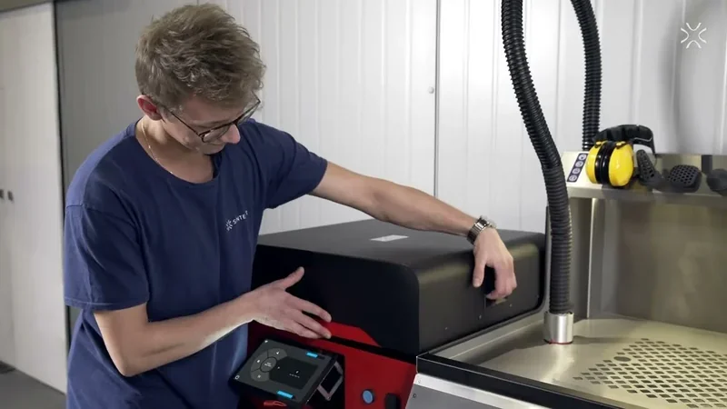 Sinterit Lisa X 3D printer operation in a workshop setting, technician adjusting controls, branded shirt visible, surrounded by equipment.