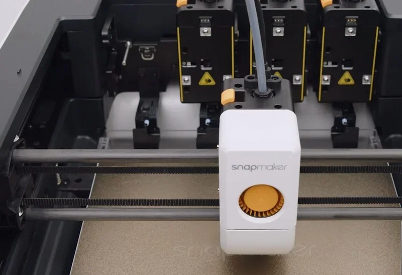 Snapmaker U1 3D printer close-up with white extruder and orange dial, printing bed below, showcasing precision parts and modular design.