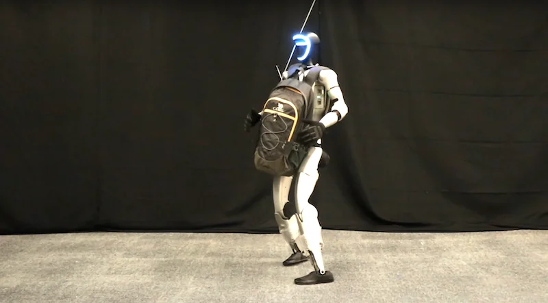 Unitree G1 robot holding a backpack, standing on a carpeted floor with a black background, showcasing its humanoid design and carrying capability.