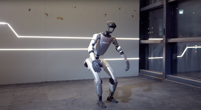 The Unitree G1 humanoid robot is pictured in a modern interior setting, standing on a gray concrete floor. Its design features a sleek, futuristic form with a predominantly metallic silver finish and black highlights on the joints. The robot is equipped with articulated limbs, a streamlined head, and a branding logo on the chest. The background includes industrial elements and illuminated lines along the wall, creating a technological ambiance.