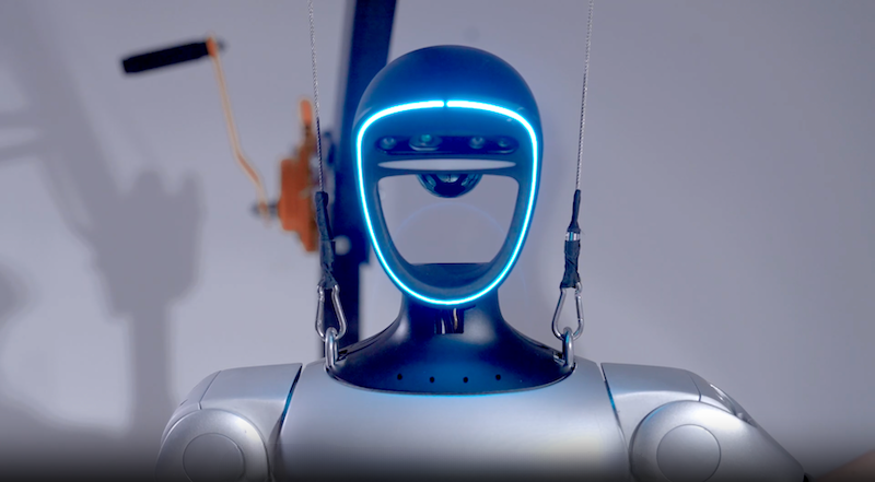 The Unitree G1 humanoid robot is featured in a laboratory setting. The robot’s head is designed with a prominent U-shaped LED display that glows blue, highlighting its sleek, futuristic design. The robot is suspended by wires for support, indicative of testing or development, with machinery visible in the blurred background that suggests an engineering environment. The robot's torso and shoulders have a metallic finish, complementing the overall advanced aesthetic.