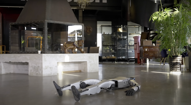 A humanoid robot from Unitree G1, lying on its back on a polished concrete floor inside a modern, well-furnished room. The robot features sleek, metallic limbs and a compact central body. It has articulated arms and legs, designed for mobility and dexterity. Surrounding the robot are various upscale interior elements including a large fireplace, decorative objects, and plant arrangements, emphasizing a blend of advanced technology within a luxurious setting.