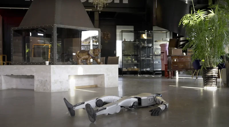 Unitree G1 robot lies on the modern indoor floor near a fireplace and furniture, surrounded by decor and plants, suggesting a tech-themed showroom.