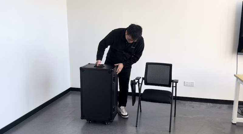 A person in a black jacket and white sneakers is handling a large black rolling case in a minimalist indoor setting, near a black chair. The scene implies preparation or transportation related to the Unitree G1, suggesting that the case may be for storing or transporting robot components or equipment. The room features a simple layout with a television screen and a table visible at the edges.