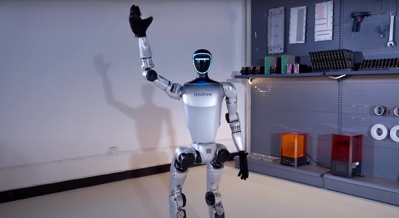 Unitree G1 robot waving in a workshop, with parts and tools neatly organized on shelves.