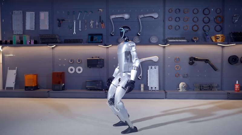 The Unitree G1 humanoid robot stands in a workshop setting, surrounded by an array of tools, electronic components, and mechanical parts on the walls and shelves. The robot, with its polished metallic surface and blue-lit face panel, is poised slightly forward, exhibiting its stability and advanced engineering. The organized workshop environment reflects its use in robotic development and testing, emphasizing the robot's purpose in controlled experimental contexts.