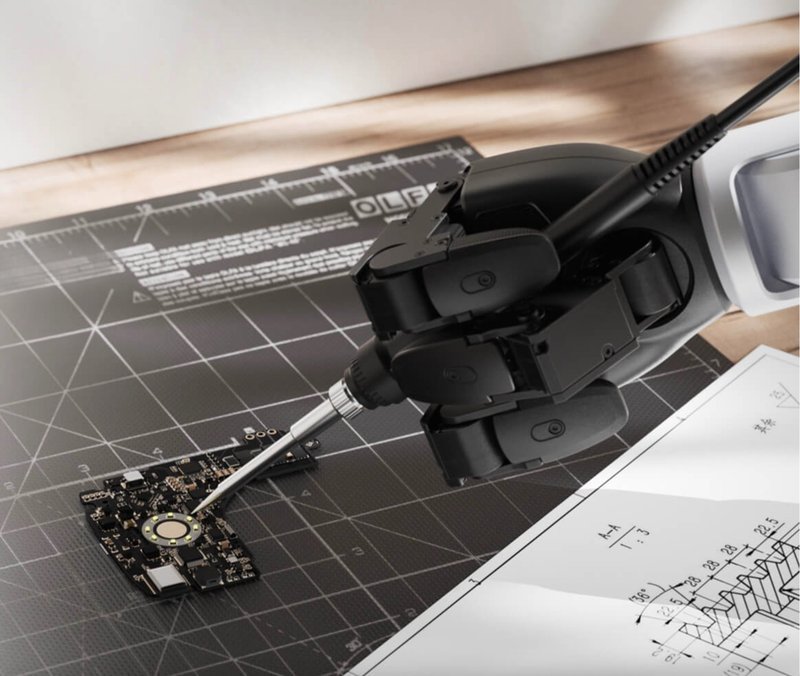 The Unitree G1's advanced robotic hand is engaged in precise electronic work, holding a soldering tool while meticulously working on a circuit board. The setting includes a cutting mat with visible measurement lines, enhancing the technical environment. Detailed imagery of the circuit board showcases multiple components, wires, and connectivity points, reflecting the robot's capability in handling delicate, intricate tasks with high precision.