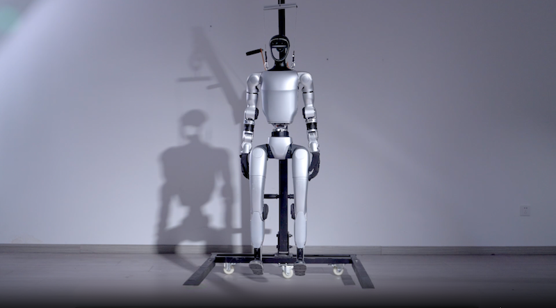 The Unitree G1 humanoid robot is positioned upright on a sleek black wheeled stand in a minimalist room with soft lighting. Its body features smooth, polished surfaces and articulated limbs. The robot's design highlights its high-tech joints and components, resembling human proportions. A prominent shadow cast on the wall behind adds depth to the scene.
