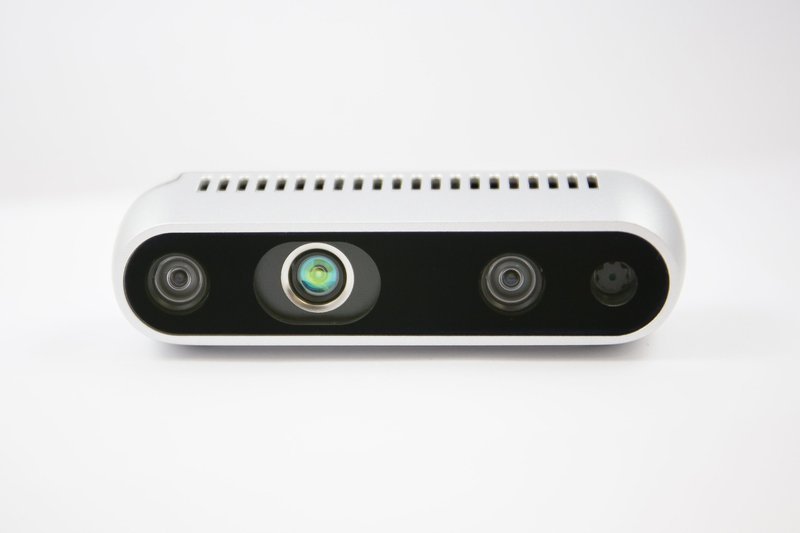 A close-up view of a sleek, modern sensor unit with three lenses aligned horizontally across its front panel. The device, characterized by a minimalist design, features a silver body with evenly spaced ventilation slits along its top. Each lens appears to be different, suggesting various functionalities such as depth sensing or imaging. The setting is a simple white background, emphasizing the unit's contemporary design.