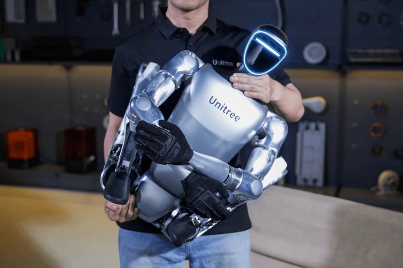 The Unitree G1 robot held by a person in a workshop setting. The robot features a metallic body with sleek, articulated arms and hands. Its face displays a luminous strip forming minimalistic facial features. The robot's torso is marked with the Unitree branding. The setting suggests a technical or robotic environment, with tools and equipment visible in the background.