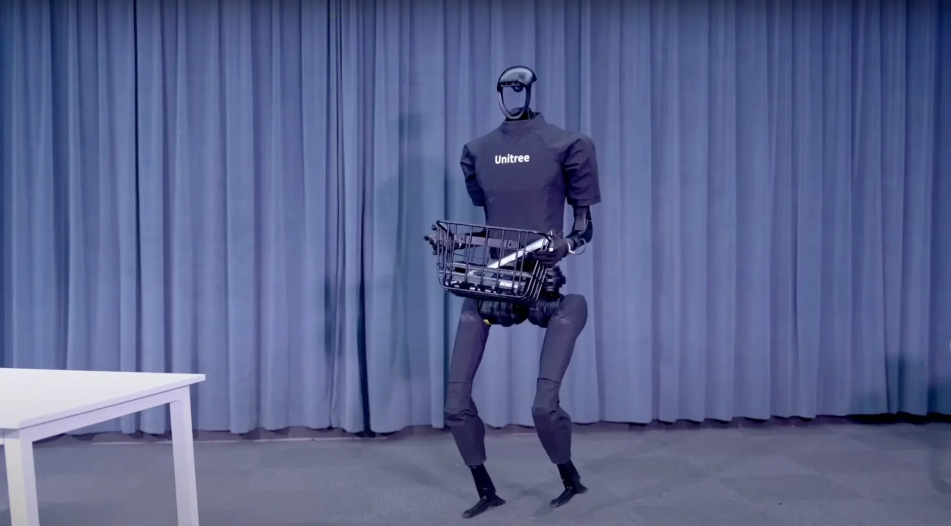 Unitree H1 humanoid robot holds a basket while standing in front of blue curtains, displaying its mobility and handling capability.