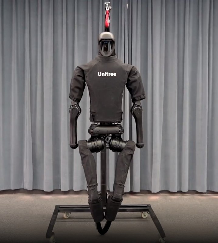 The Unitree H1 humanoid robot is displayed hanging from a support in front of a gray curtain. The robot features articulated arms and legs with joint segmentation, and its overall structure resembles a human form with a distinct helmet-like head. The branding "Unitree" is prominently visible on the chest area of the robot. The setting suggests a controlled indoor environment, possibly for demonstration or testing.