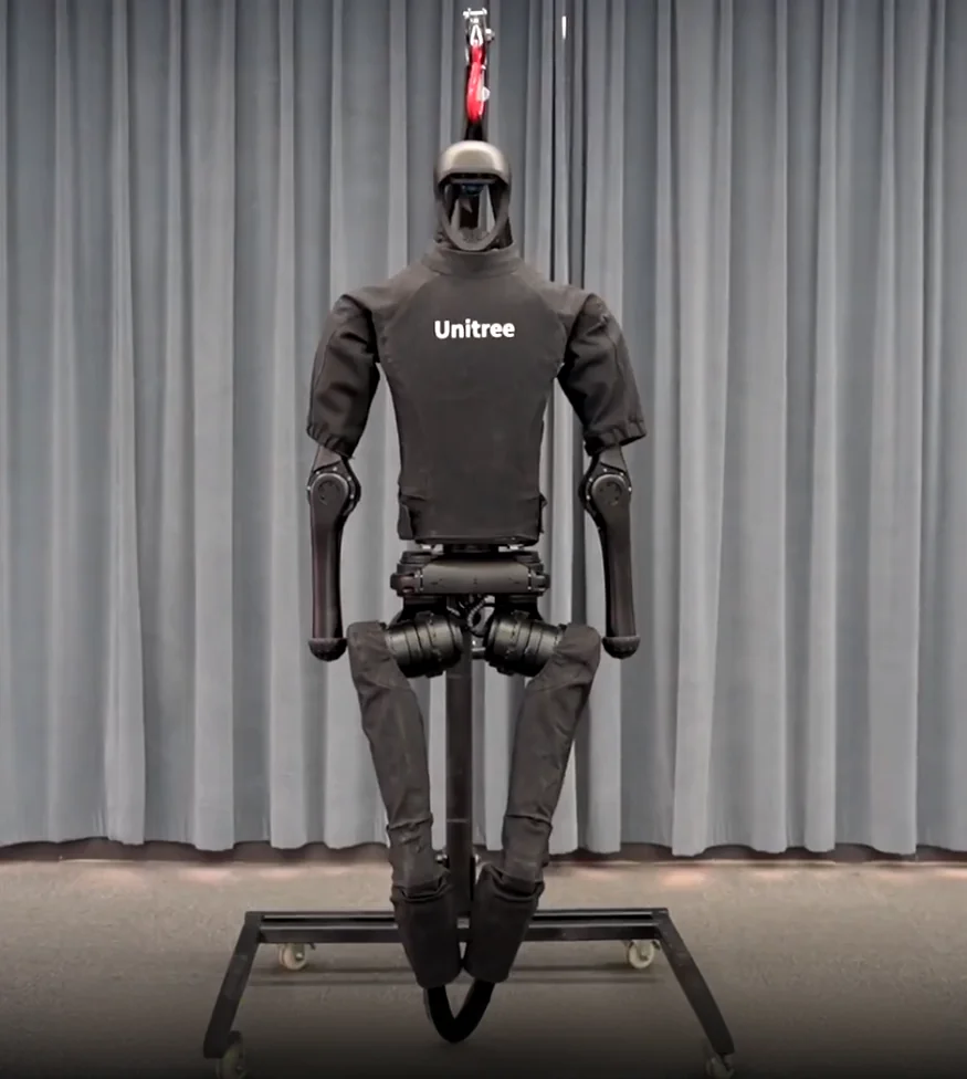 Unitree H1 humanoid robot on a wheeled stand against a curtain backdrop. Branding visible on the torso.