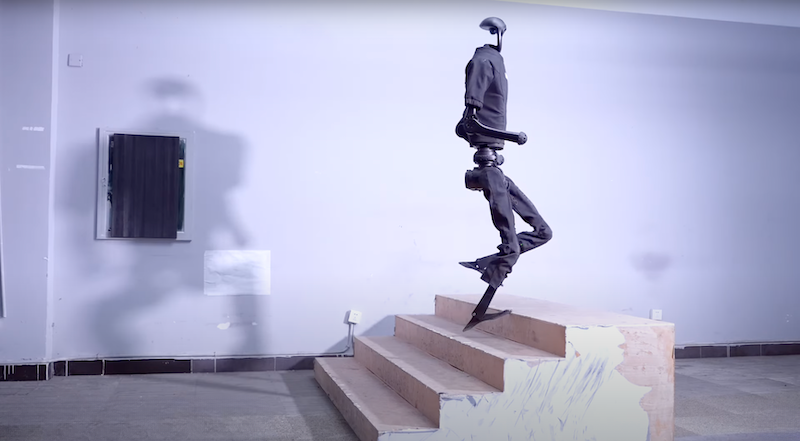 The Unitree H1 humanoid robot is seen in motion, actively ascending a set of wooden stairs within an indoor environment. The robot's design is streamlined with articulated joints simulating a human walking motion. The space appears to be a workshop or testing area with minimal decor, highlighting the robot's balance and stability as it navigates the steps.