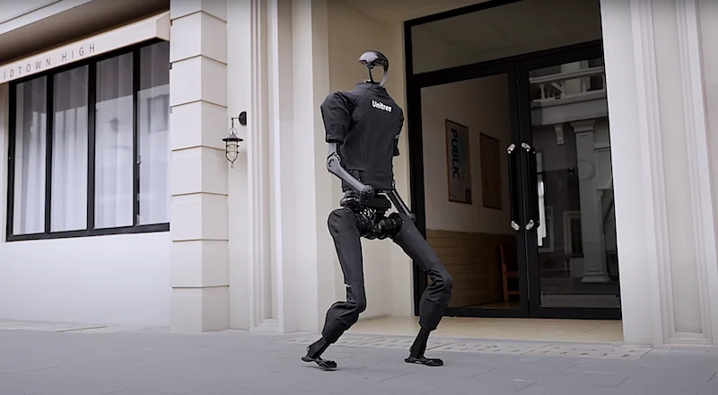 Unitree H1 robot in black, humanoid form, standing in front of glass entrance, wearing "Unitree" branded shirt outside Midtown High building.