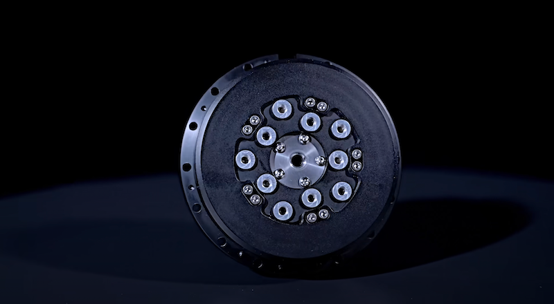 A close-up of a circular black component, possibly a motor or actuator part used in the Unitree H1 robot. The component has multiple evenly spaced screws and circular elements on its face, indicating its mechanical complexity and precision. The setting is against a dark background, emphasizing its intricate design and likely critical role in the robot's movement.